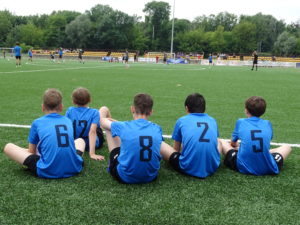 młodzi piłkarze w granatowych strojach siedzą na murawie boiska i oglądają toczący się mecz.